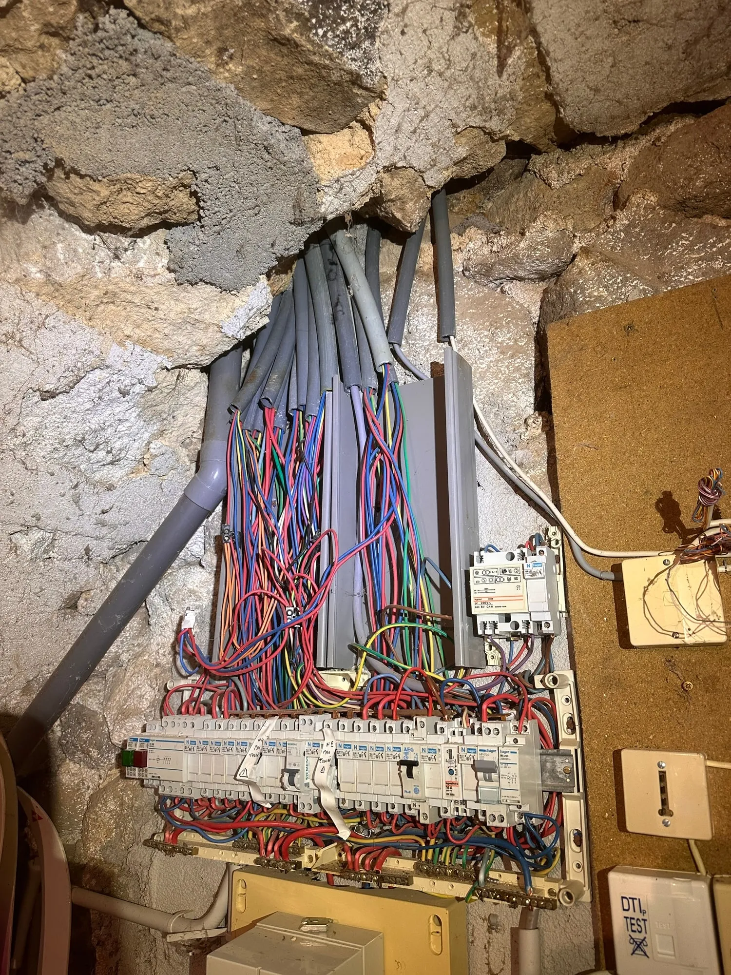Installation électrique vétuste à Ailhon : tableau ouvert et arrivée des câbles sur un mur en pierre.